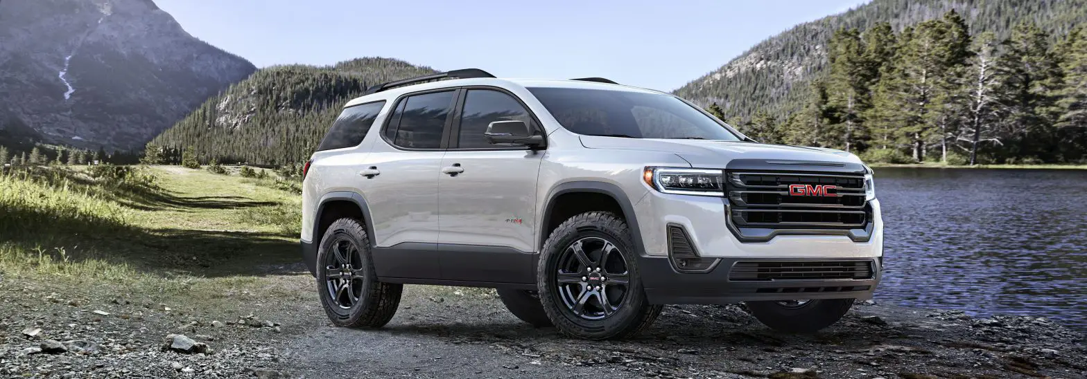 Front driver 3/4 view of 2023 GMC Acadia AT4 in Summit White with mountains and lake in background.