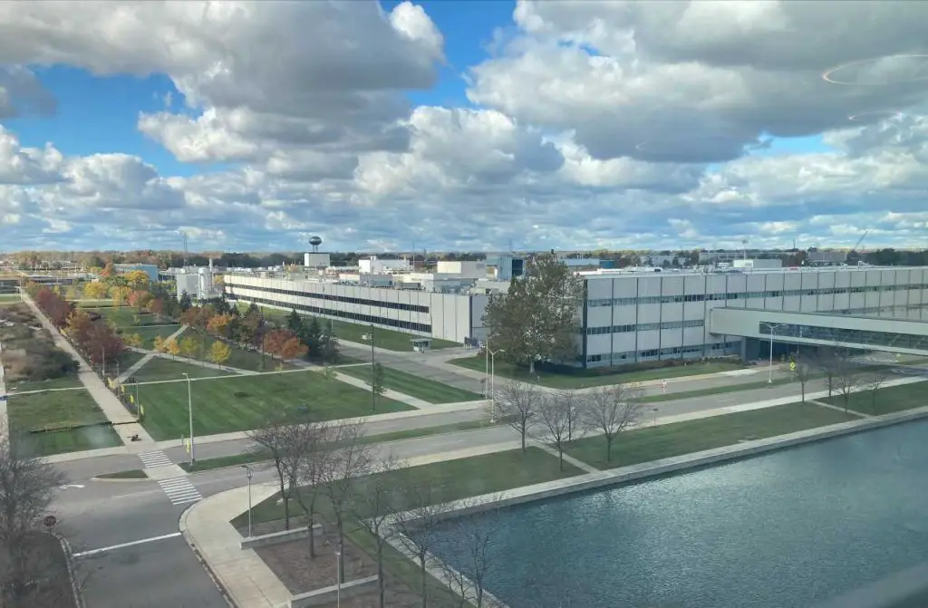 GM Lays Off 1,000 Workers, Including the Brake Quality Guy - What Could Go Wrong? 2 Aerial view of GM's Warren Technical Center on a partly cloudy day, showing a large industrial building complex, landscaped walkways, and a nearby water feature.