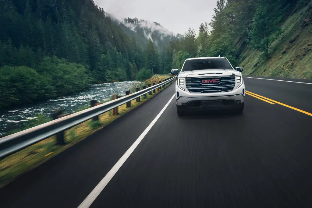 Mexican Tariffs Could Drive Chevy Silverado & GMC Sierra Prices Up $13,750 3 A GMC Sierra driving along a scenic road by a river, surrounded by dense forest and mountains, highlighting its rugged yet refined design.