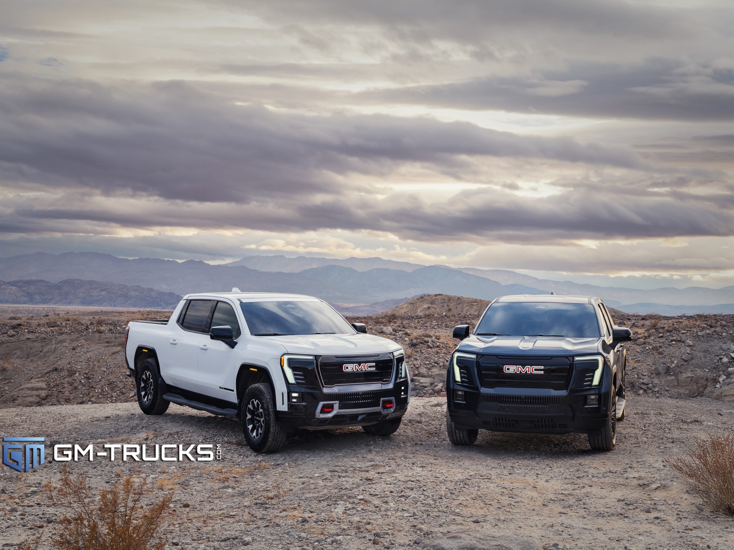 Still Don’t Trust EV Trucks? The 2026 GMC Sierra EV Is Built to Change That With A Lower Price & More Trims 2 A white 2026 GMC Sierra EV AT4 and a black Elevation trim parked side by side on rocky terrain under a cloudy desert sky, highlighting their distinct features.