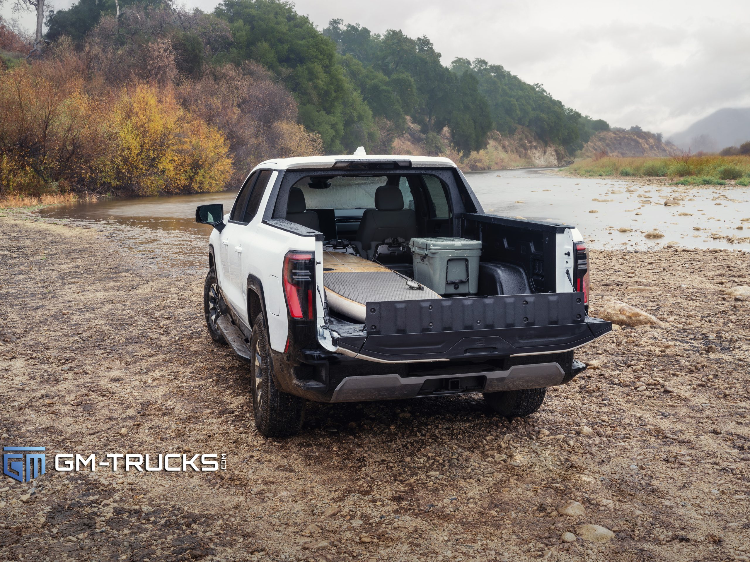 Still Don’t Trust EV Trucks? The 2026 GMC Sierra EV Is Built to Change That With A Lower Price & More Trims 3 A rear view of a white 2026 GMC Sierra EV AT4 parked near a riverbank with its MultiPro MidGate and tailgate open, showing extended bed space and gear storage.