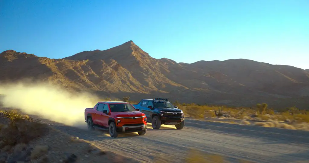A Chevrolet Silverado EV Trail Boss and a Silverado EV ZR2 race truck kicking up dust while driving side by side through the desert, showcasing off-road capability.