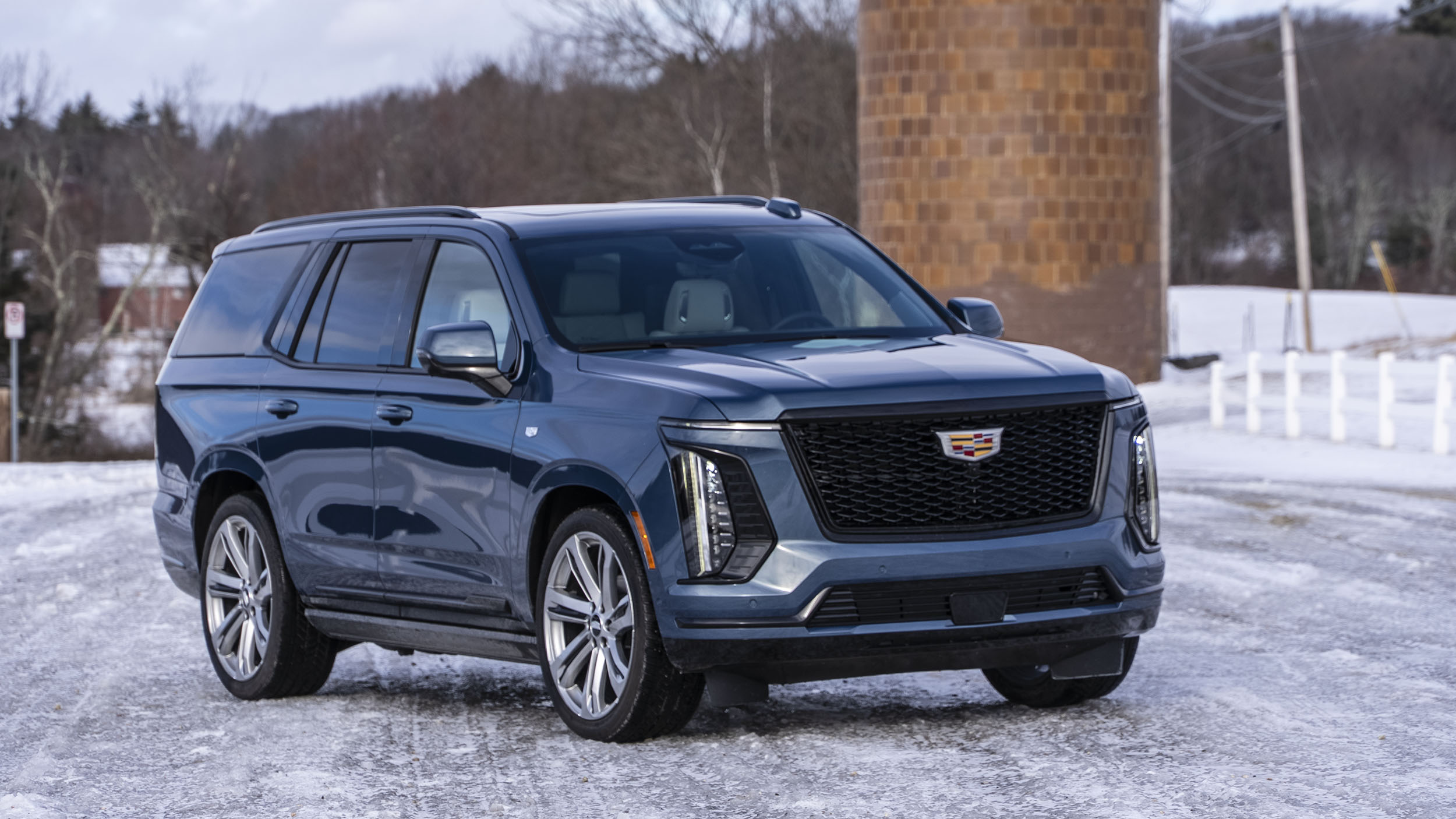 Power Open Doors, Night Vision & 55-Inch Displays: 5 Wild Tech Features on the 2026 Escalade 2 A front three-quarter view of a dark blue 2026 Cadillac Escalade Sport Platinum parked on a snowy road with a rustic wooden silo and bare trees in the background.