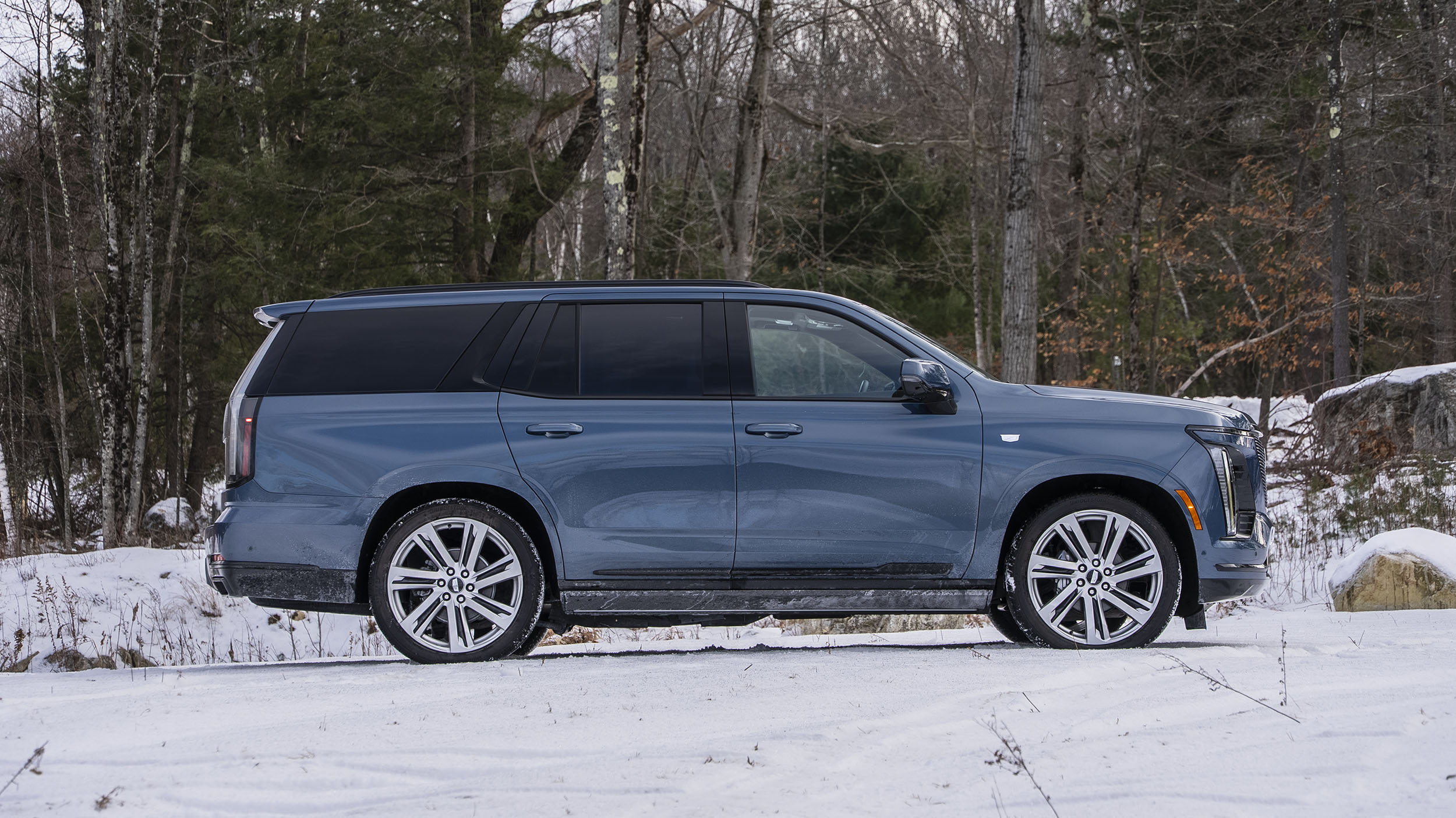 Power Open Doors, Night Vision & 55-Inch Displays: 5 Wild Tech Features on the 2026 Escalade 7 A full side profile shot of a dark blue 2026 Cadillac Escalade Sport Platinum parked on a snowy road with a forest of bare trees in the background.