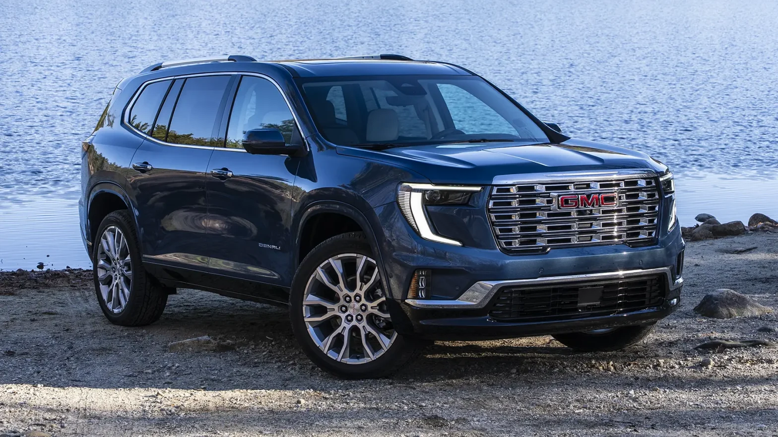 A dark blue GMC Acadia Denali parked on a dirt shoreline next to a body of water, representing the premium trim that receives standard Super Cruise for the 2027 model year.