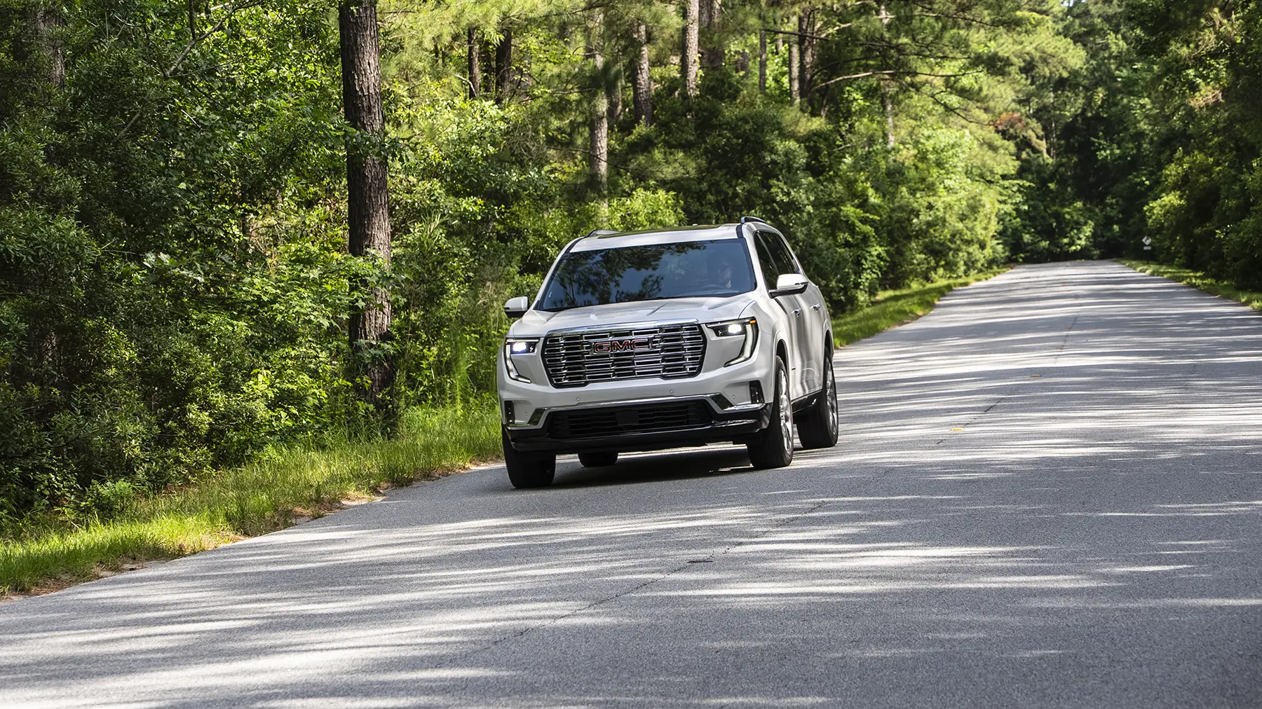 2027 GMC Acadia Updates: Standard Denali Super Cruise, Interior Camera & AT4 Reserve 4 A white GMC Acadia Denali driving on a paved, tree-lined road, highlighting the premium trim that now features standard Super Cruise.