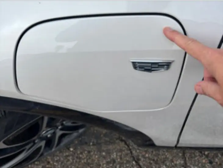 Cadillac To Optiq Owners: That Emblem Is Not A Button, Stop Pushing It 3 A close-up photograph showing a finger pressing the upper right-hand corner of a flush-fitting white Cadillac Optiq charge port door, completely avoiding the Cadillac emblem right below it.