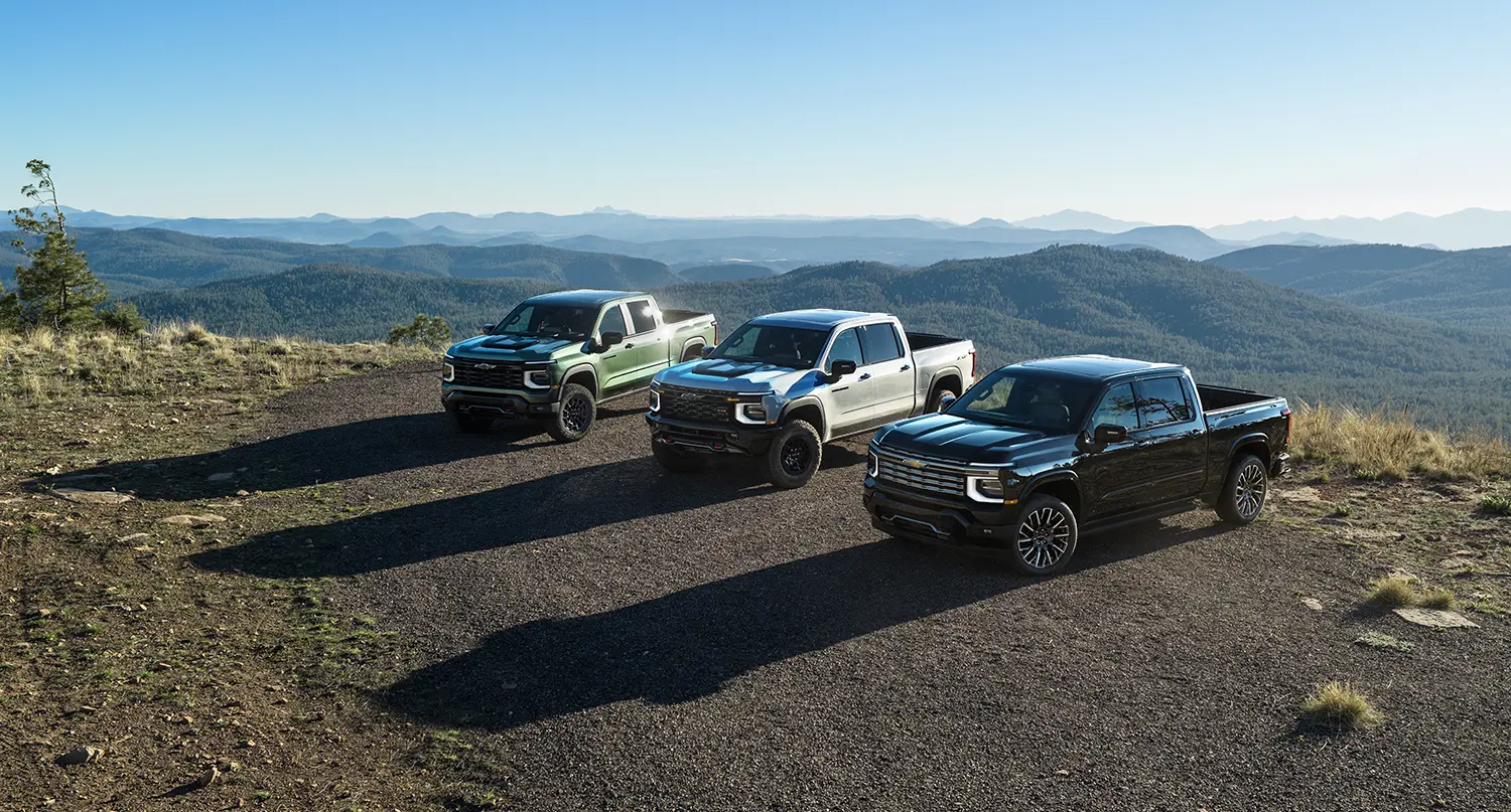 Three 2027 Chevrolet Silverado 1500 trucks parked on a mountain overlook: a green Trail Boss, a silver ZR2, and a black High Country.