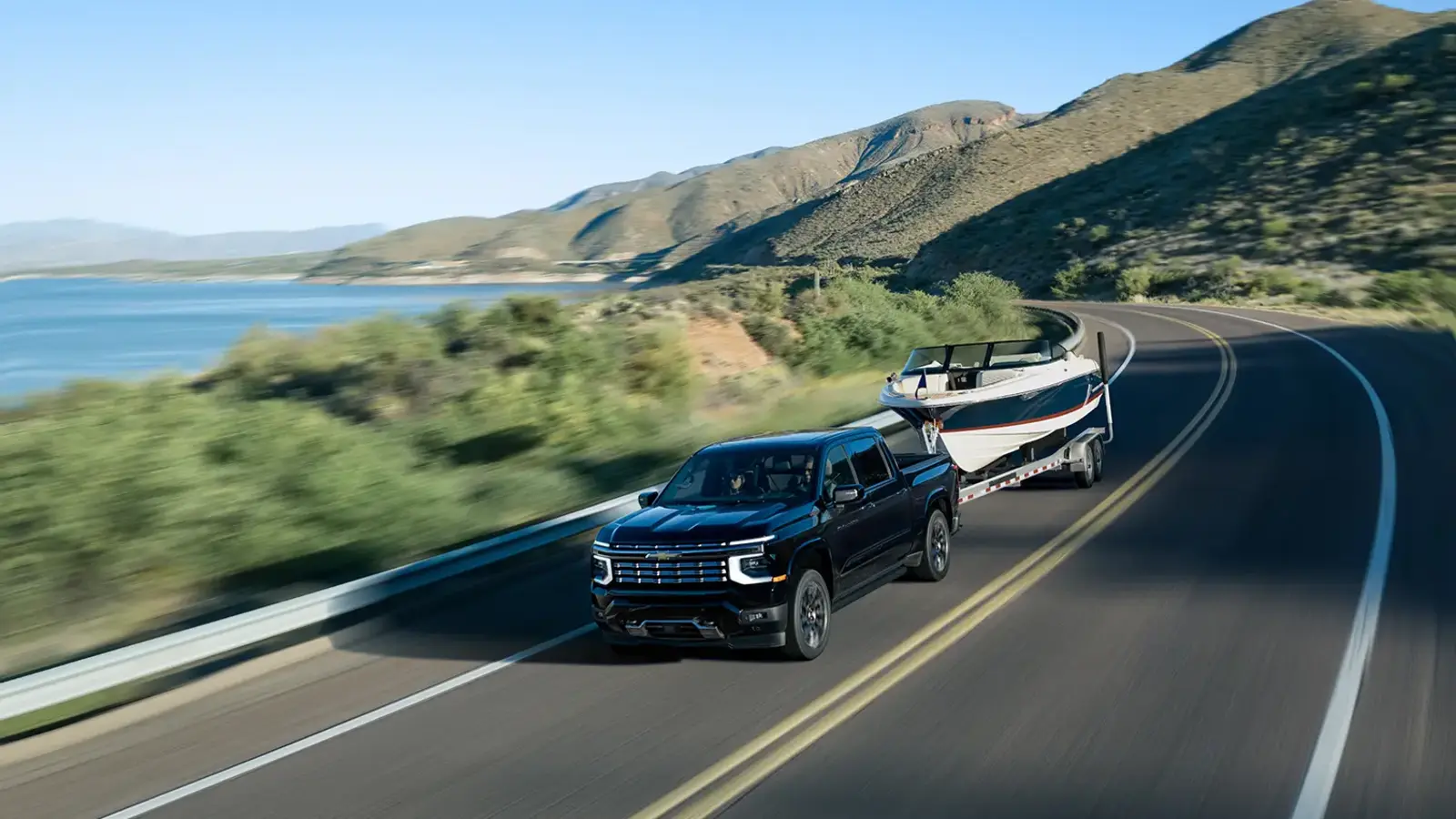 L76 and L78 Return: GM's New V8s Get Muscle Car Codes 3 Dark blue 2027 Chevrolet Silverado 1500 High Country pickup truck towing a boat on a winding highway beside a lake