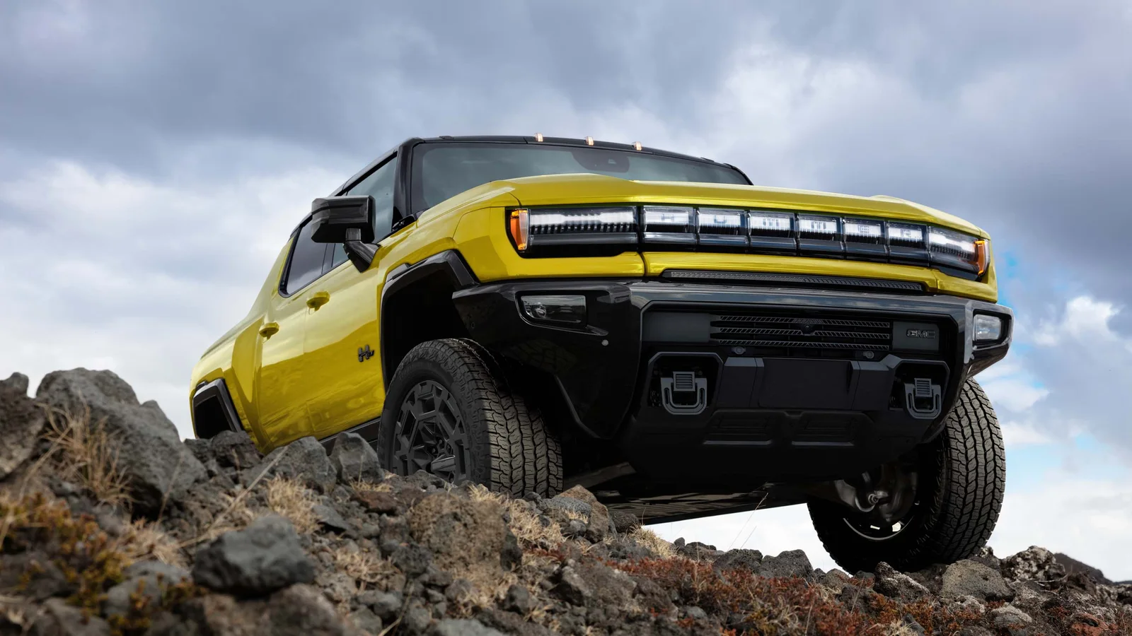 Hummer Turns 25 and GMC Brought the H2's Yellow Back 5 Yellow 2027 GMC HUMMER EV Pickup climbing over rocky terrain under a cloudy sky