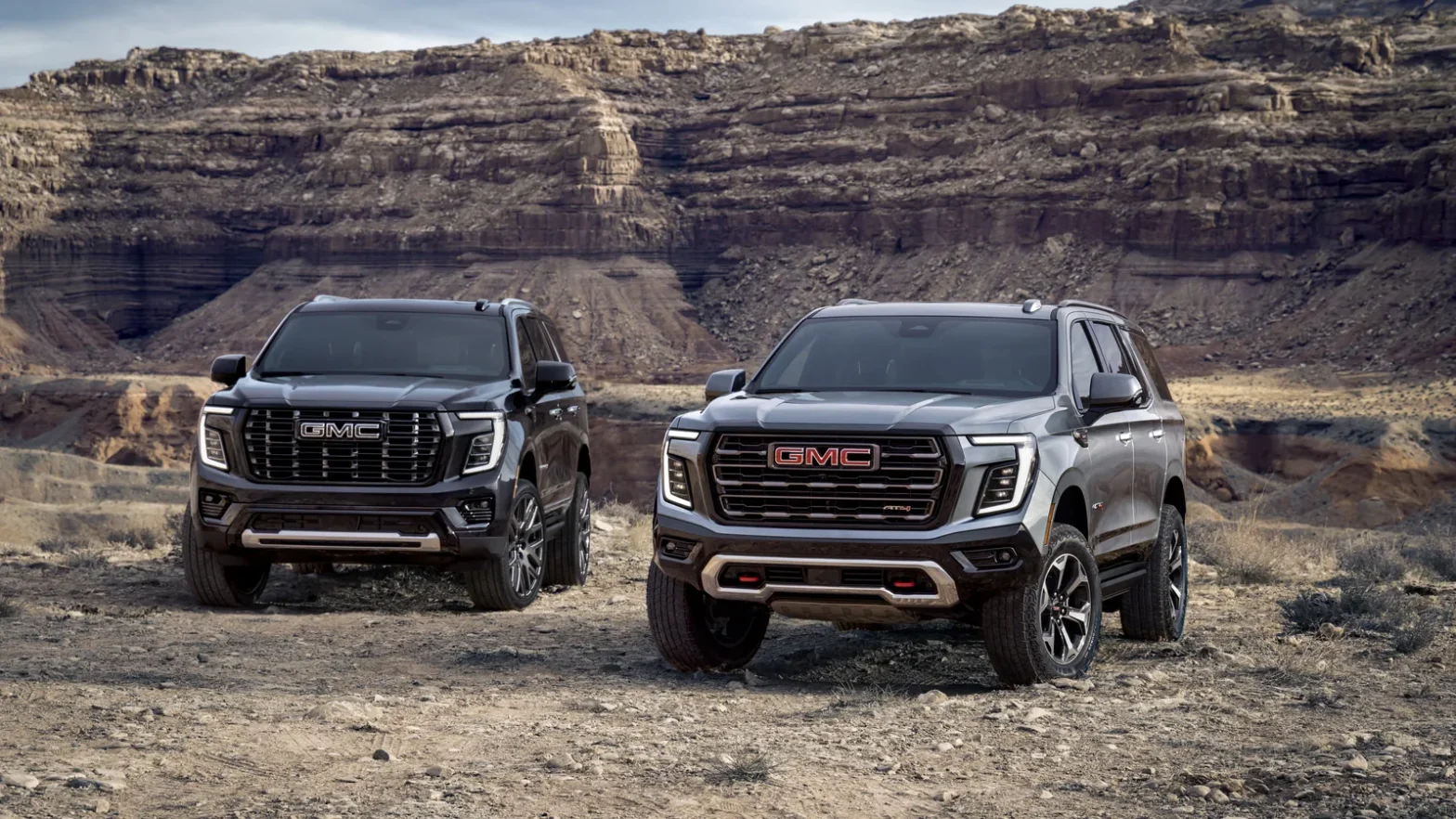 2027 GMC Yukon Denali and Yukon AT4 parked on desert terrain in front of a rocky canyon wall