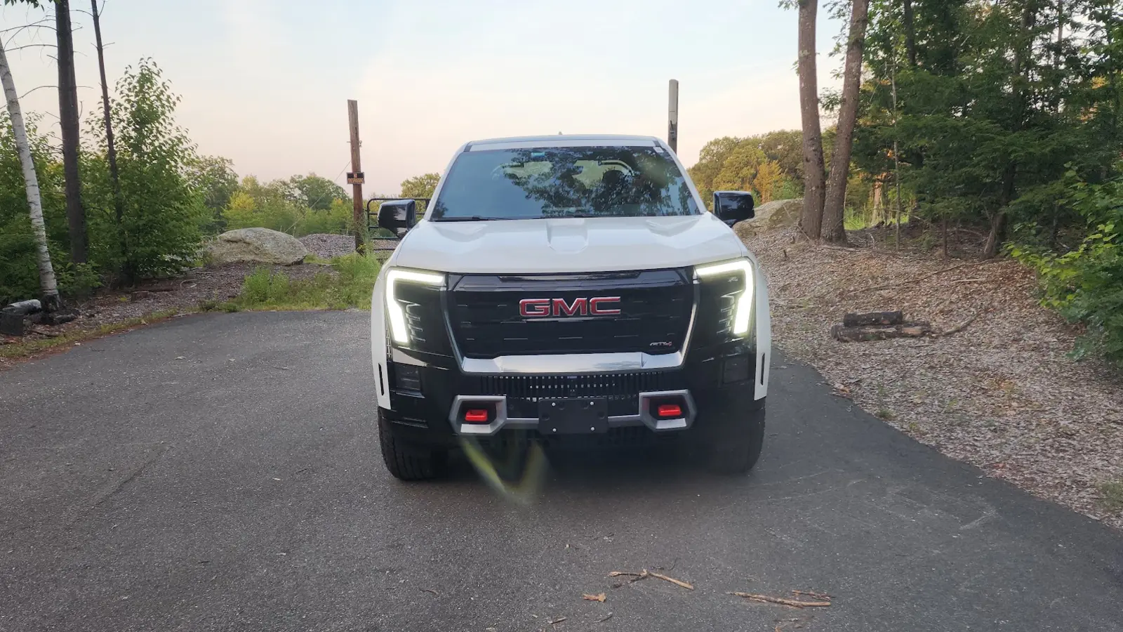 GM Ends Mandatory Vehicle-Wide Programming on EVs 2 White GMC Sierra EV AT4 electric pickup facing forward on a wooded driveway with its LED headlights on