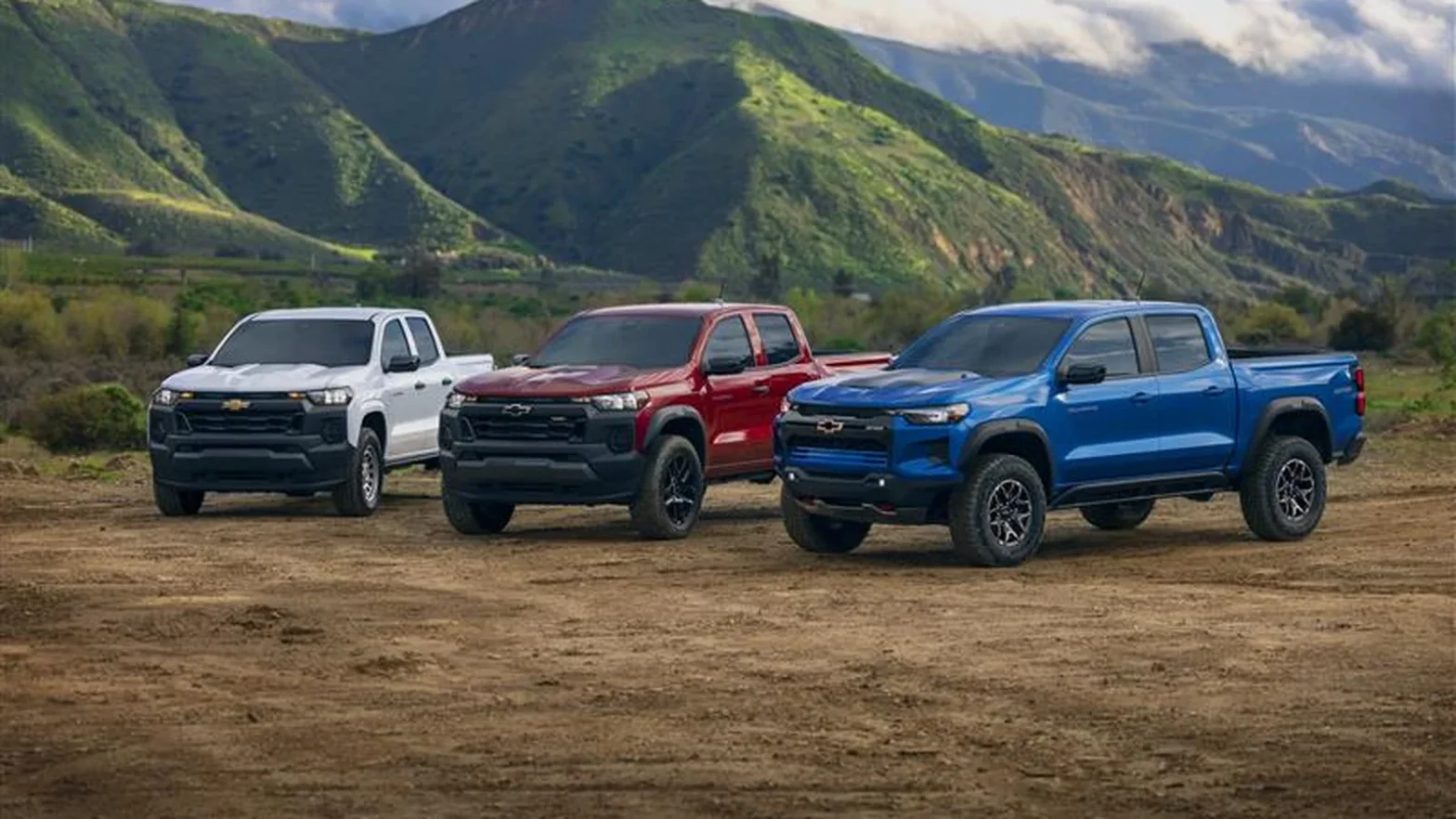 GM Opens 2027 Chevrolet Colorado Order Books - Here's What's New 1 Three third-generation Chevrolet Colorado pickup trucks parked on dirt in front of green mountains, in white, red, and blue