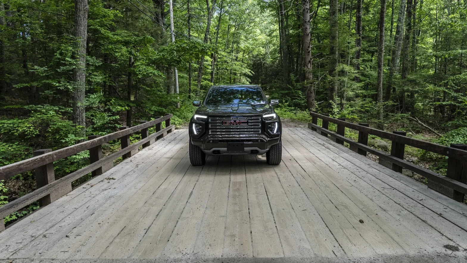 Front view of a dark GMC Canyon pickup truck crossing a wooden plank bridge surrounded by green forest
