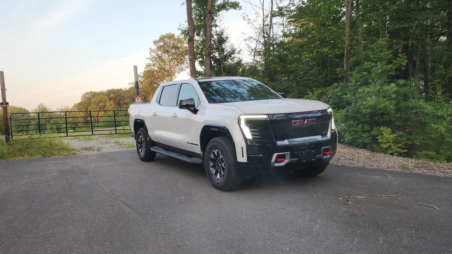 White GMC Sierra EV AT4 crew cab electric pickup parked on a paved overlook surrounded by trees at dusk